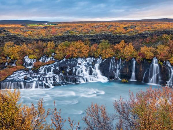 Numerous waterfalls cascade into a milky blue river, surrounded by vibrant autumn foliage.