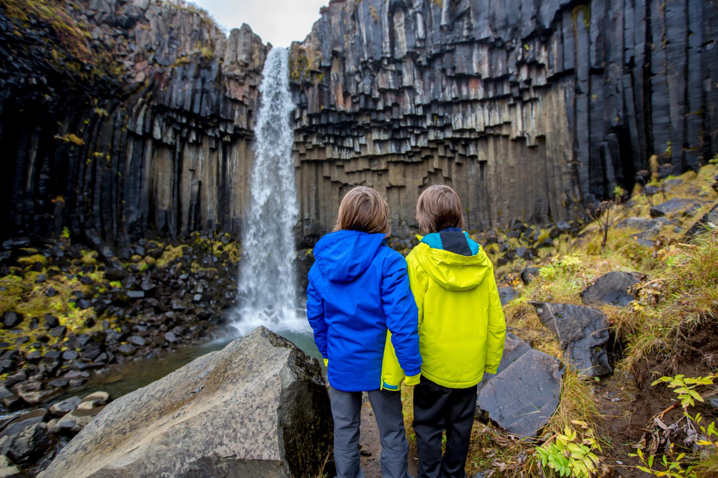 Two people in bright jackets stand with their backs to the viewer, looking at a waterfall cascading over dark columnar basalt cliffs.