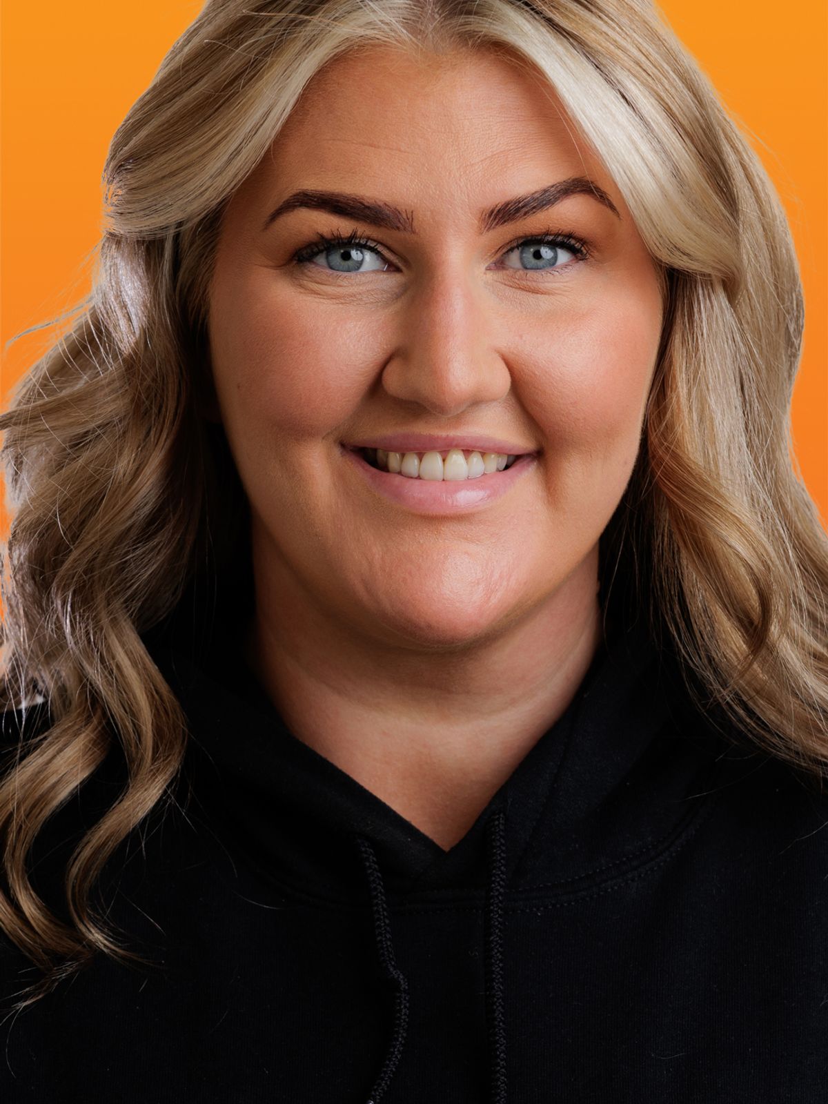 une femme en sweat à capuche noir sourit devant un fond orange.