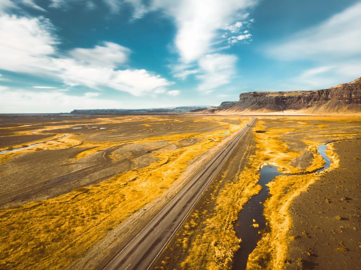 Aerial view of a long road through a vast golden landscape with a winding river and cliffs under a blue sky.