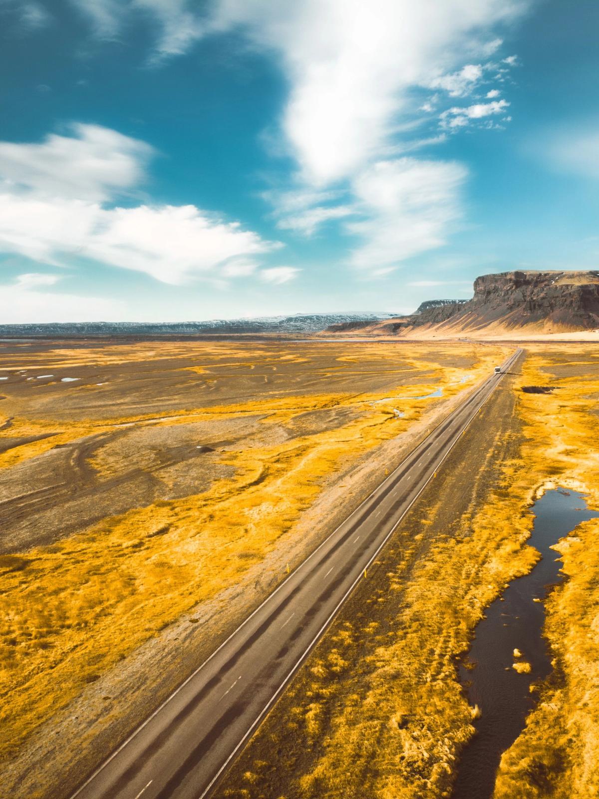 Aerial view of a long road through a vast golden landscape with a winding river and cliffs under a blue sky.