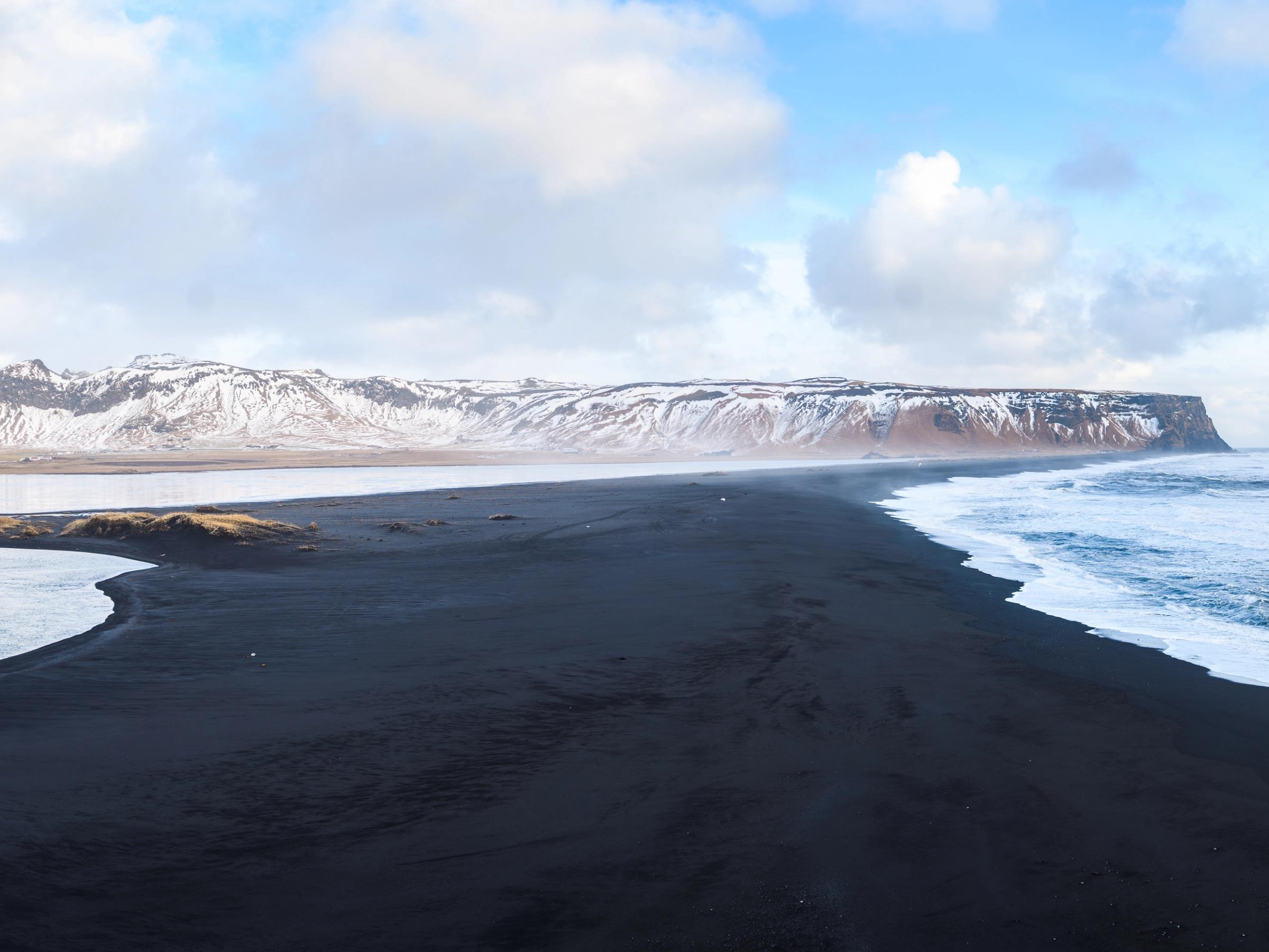 Black sand beach with ocean waves, a river, and snow-capped mountains.