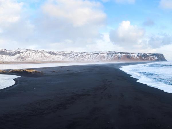 Playa de arena negra con olas del océano, un río y montañas nevadas.