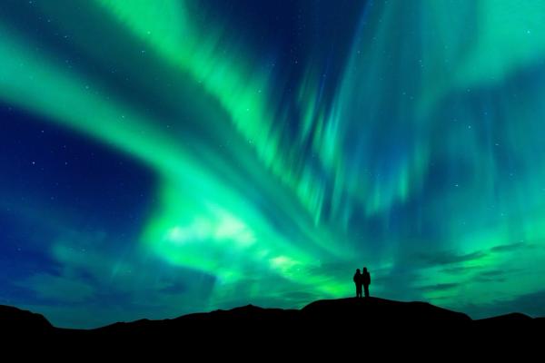 two people are standing on top of a hill watching the aurora borealis .