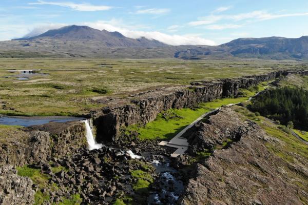 An aerial view of Thingvellir National Park in Iceland, showing a deep rocky fissure with a waterfall, green plains, and distant mountains.