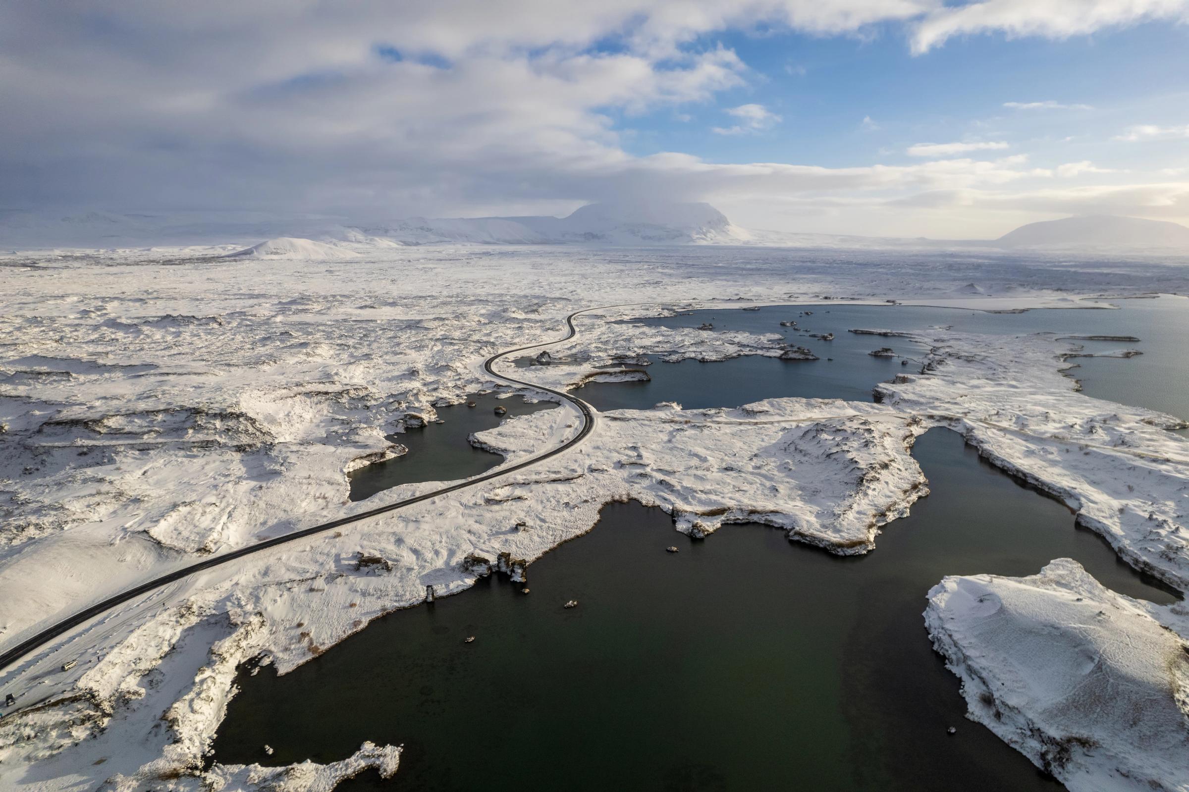 Aerial view of a winding road through a snow-covered landscape with dark lakes and distant mountains.