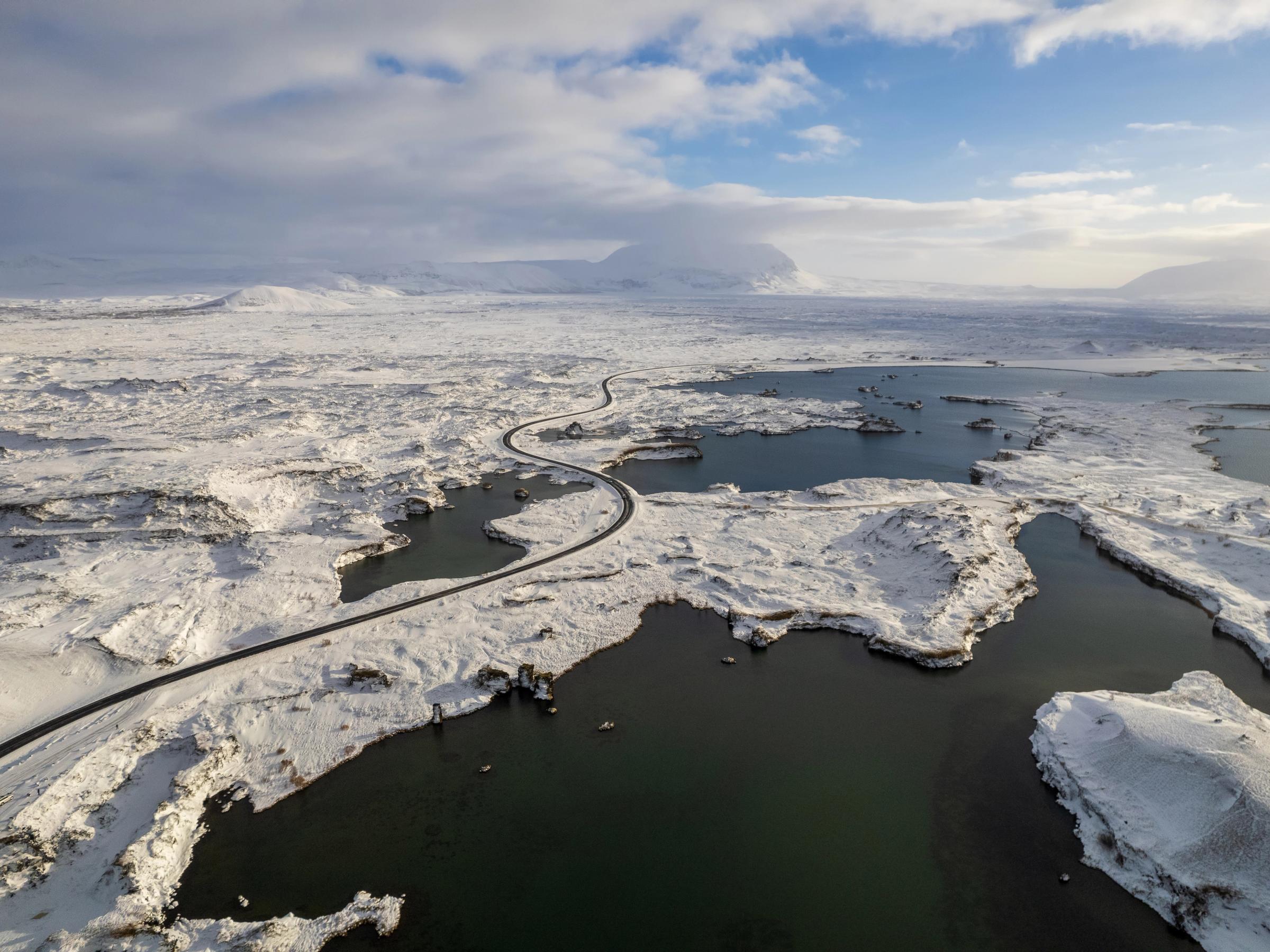 Aerial view of a winding road through a snow-covered landscape with dark lakes and distant mountains.