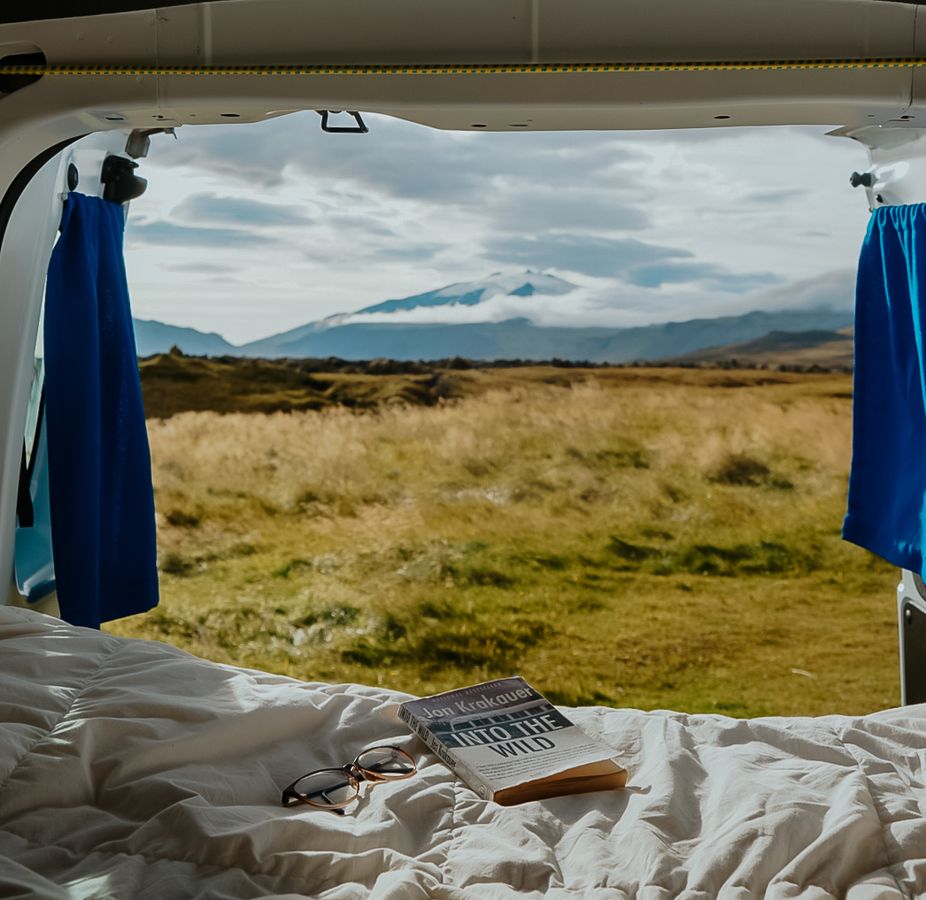 Buch und Brille auf der Bettdecke, dahinter offene Türen mit Blick auf Wiesen und Berge