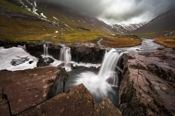 Long-exposure image of waterfalls cascading over dark rocks in a rugged mountain valley with patchy snow and a cloudy sky.