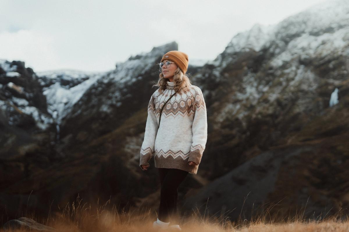 a woman in a sweater and hat is standing in a field in front of a mountain .