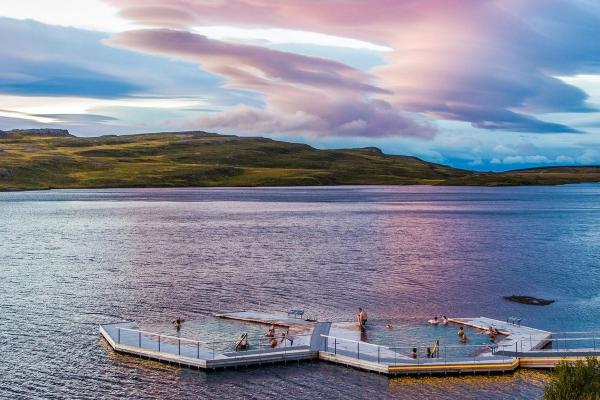 People relax in floating pools on a lake, surrounded by green hills under a dramatic pink and purple sky.
