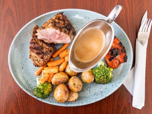 Carne asada con papas asadas, zanahorias, brócoli y una salsera en un plato, con cubiertos al lado.