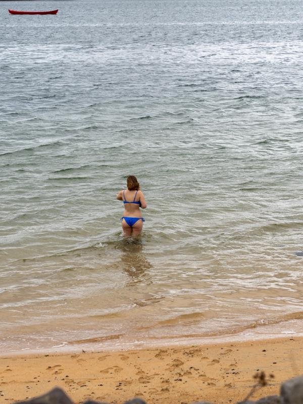 A woman in a blue bikini stands in shallow water at a sandy beach with a red boat in the distance.