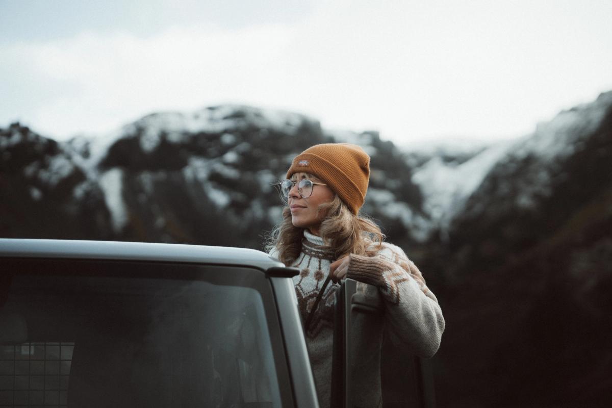 Woman in a beanie and glasses leans on a car, looking at snowy mountains.