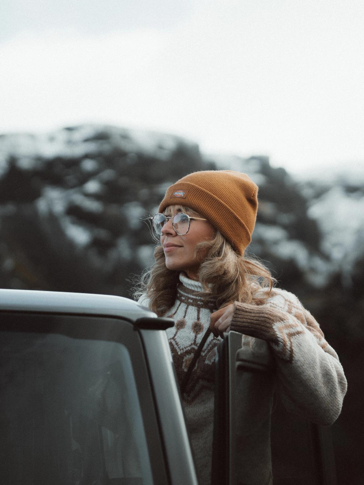 Woman in a beanie and glasses leans on a car, looking at snowy mountains.