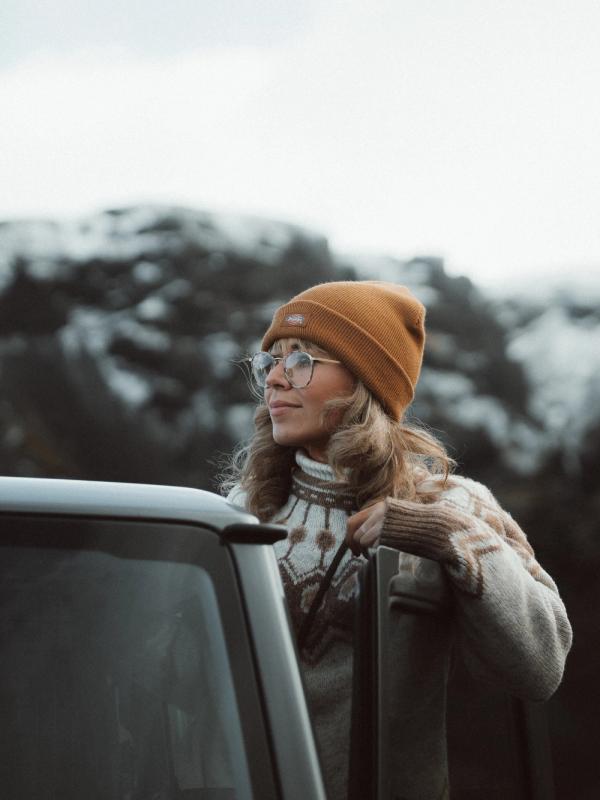Woman in a beanie and glasses leans on a car, looking at snowy mountains.