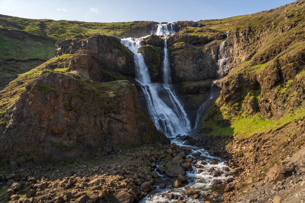 A multi-tiered waterfall cascades down a rocky, green hillside into a rocky stream.