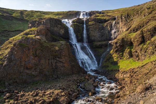 A multi-tiered waterfall cascades down a rocky, green hillside into a rocky stream.
