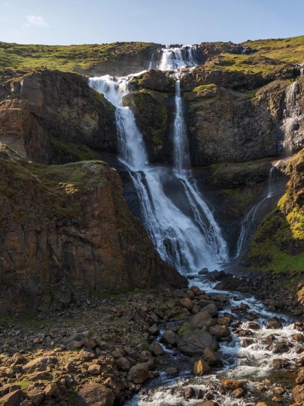 A multi-tiered waterfall cascades down a rocky, green hillside into a rocky stream.