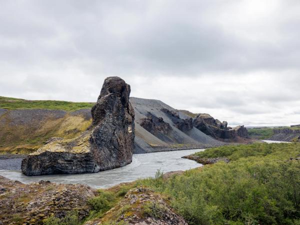 A prominent dark rock formation stands in a river flowing through a valley with green and rocky slopes under an overcast sky.