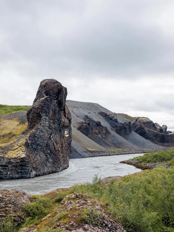 A river flows past a tall rock formation amidst steep, green and rocky banks under an overcast sky.