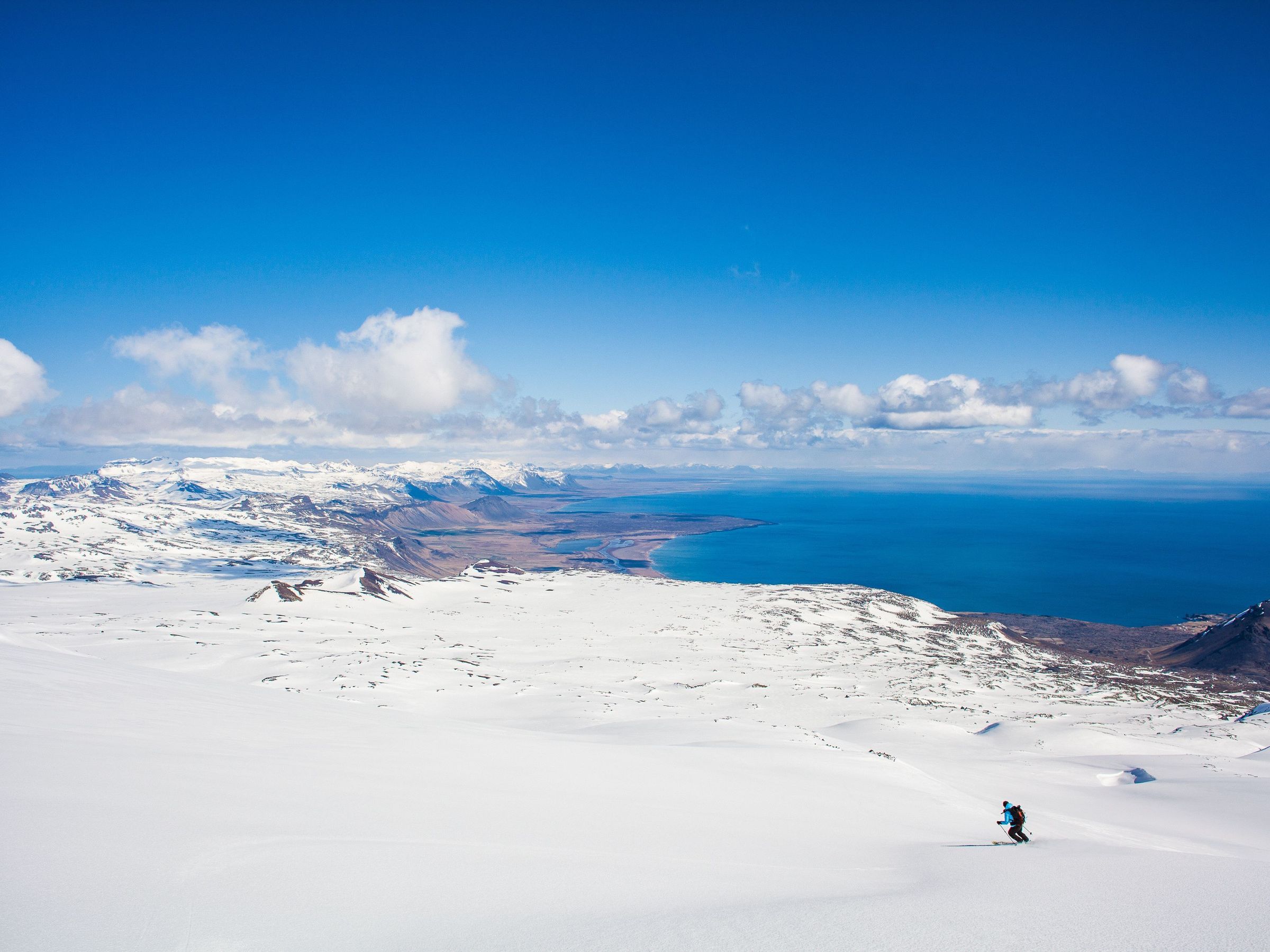 Un esquiador desciende una inmensa montaña nevada con vistas a un océano azul bajo un cielo despejado.