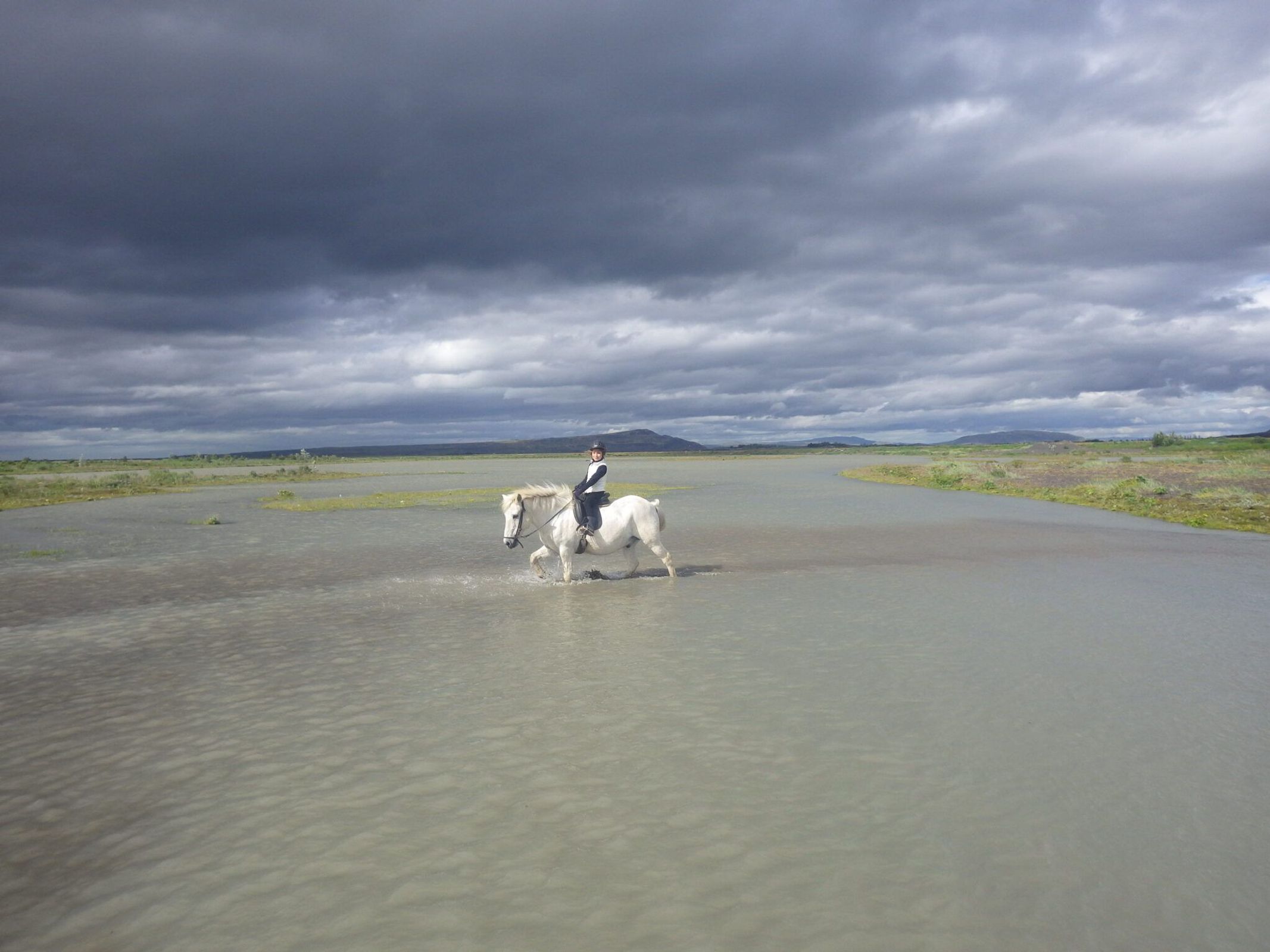 Un jinete sobre un caballo blanco cruza aguas poco profundas bajo un cielo dramático y nublado, en un paisaje llano y abierto.