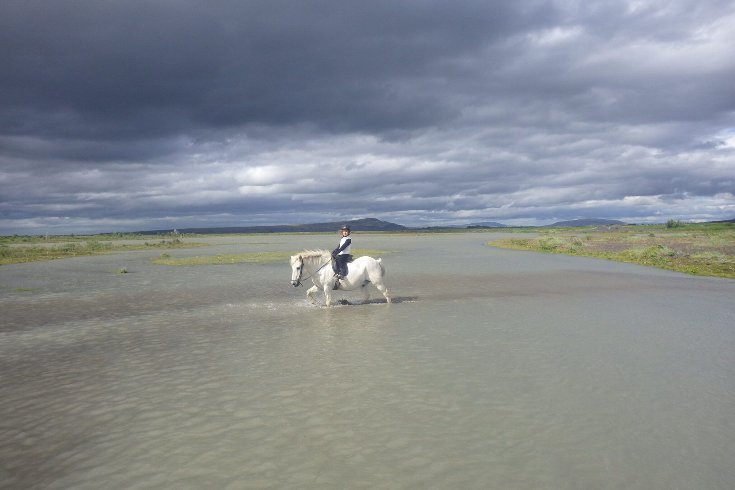 Un jinete sobre un caballo blanco cruza aguas poco profundas bajo un cielo dramático y nublado, en un paisaje llano y abierto.