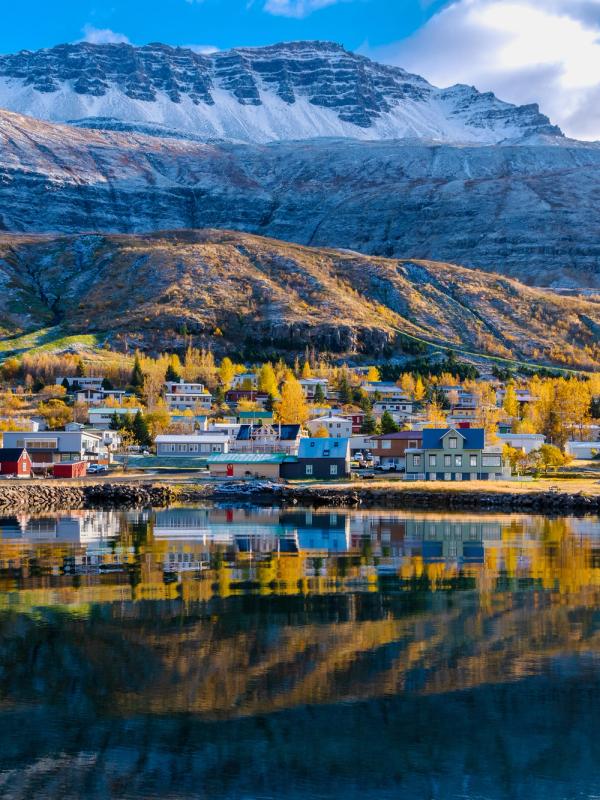 Colorful waterfront village with autumn trees and snow-capped mountains reflecting in the water.