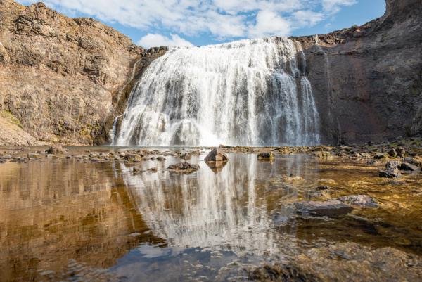 A wide waterfall cascades over a rocky cliff into a reflective pool under a blue sky.