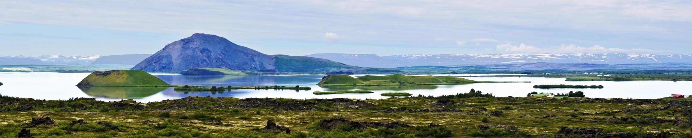 Panoramic view of Lake Myvatn with green pseudocraters, a large dark volcanic mountain, and distant snow-capped peaks.