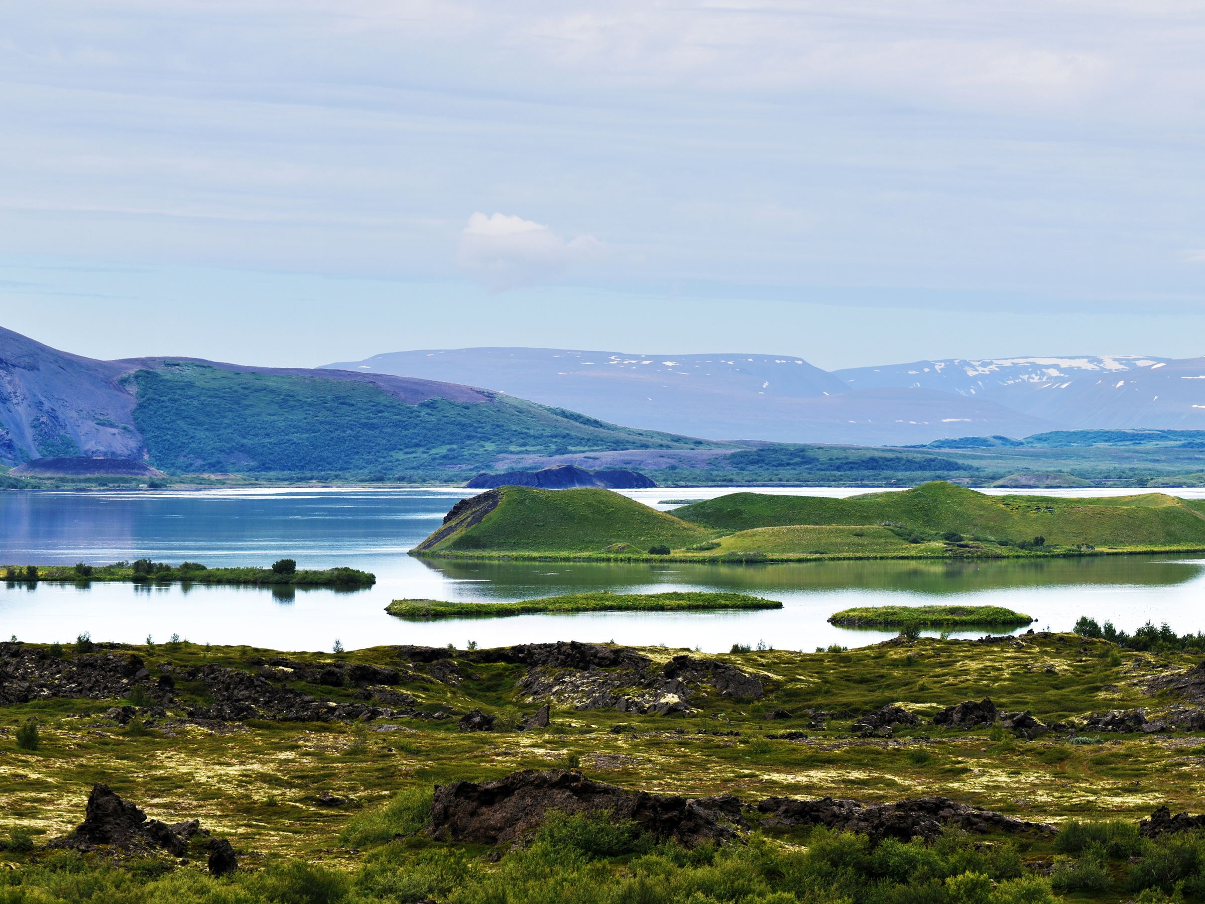 Panoramic view of a volcanic lake with pseudocraters, a large conical mountain, and distant snow-capped peaks.