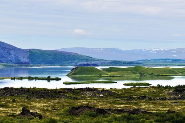 Panoramic view of Lake Myvatn with green pseudocraters, a large dark volcanic mountain, and distant snow-capped peaks.