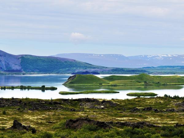 Vista panorámica de un lago volcánico con pseudocráteres, una gran montaña cónica y picos lejanos cubiertos de nieve.