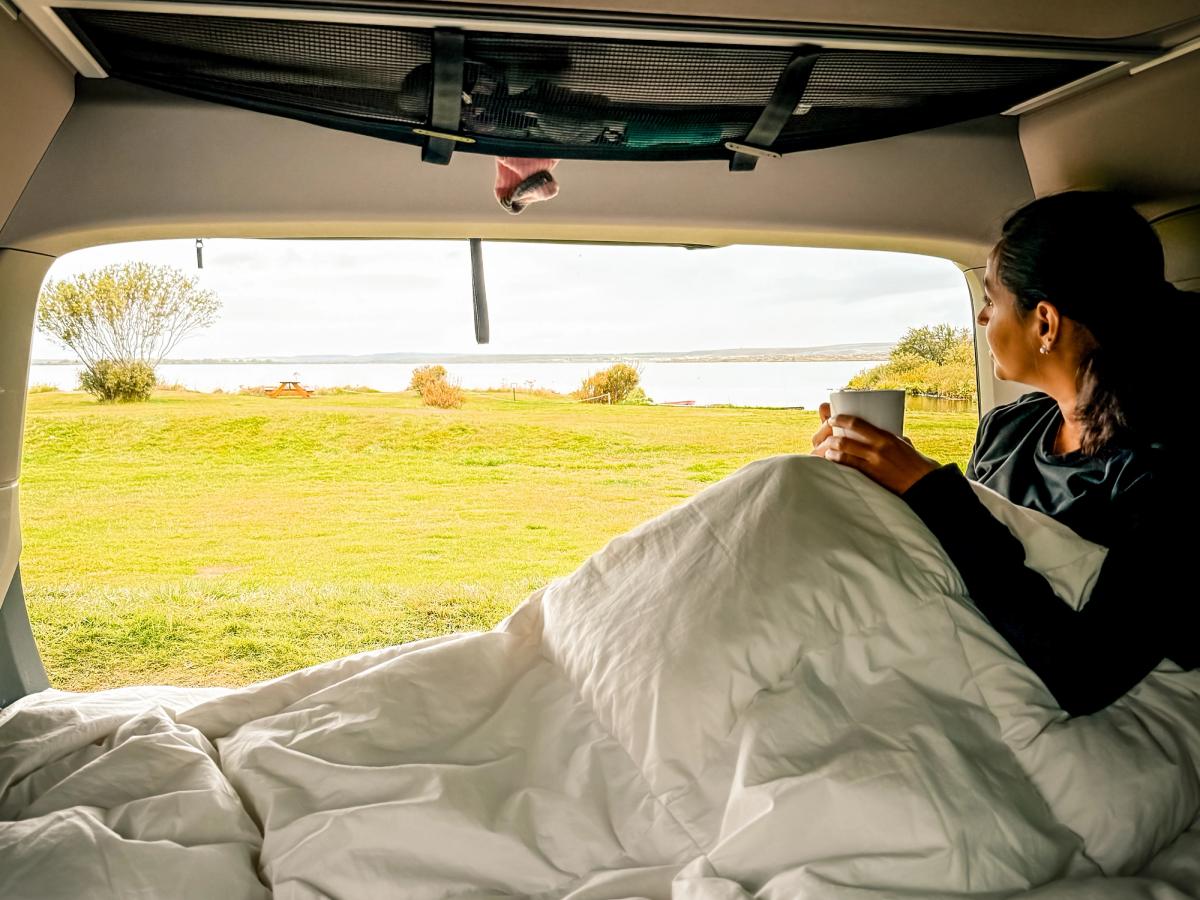 Vue de la benne depuis le hayon Personne assise sur le lit sous une couette, une tasse à la main, regardant par le hayon ouvert