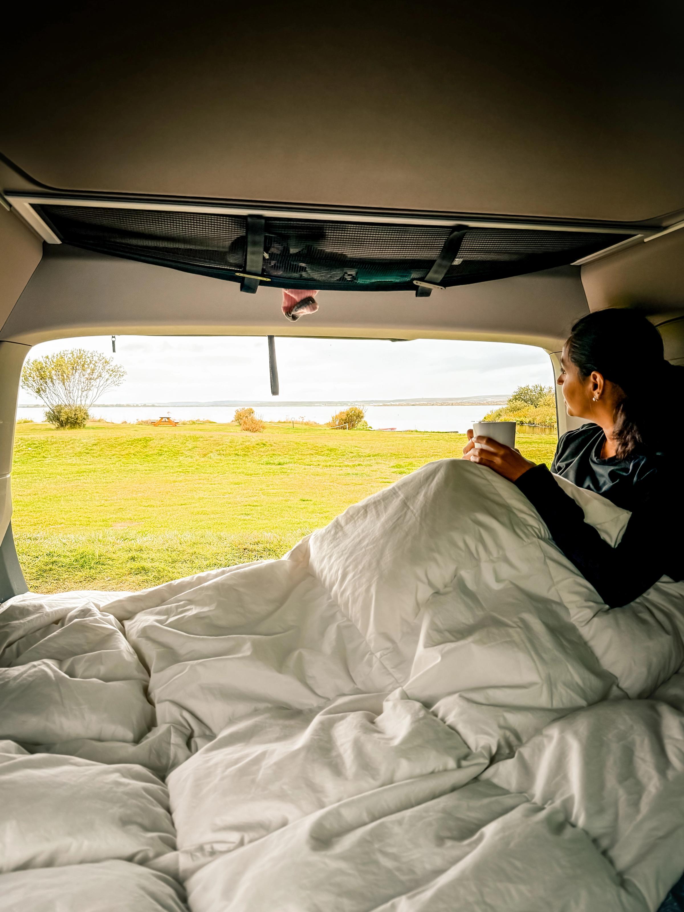 Vue de la benne depuis le hayon Personne assise sur le lit sous une couette, une tasse à la main, regardant par le hayon ouvert
