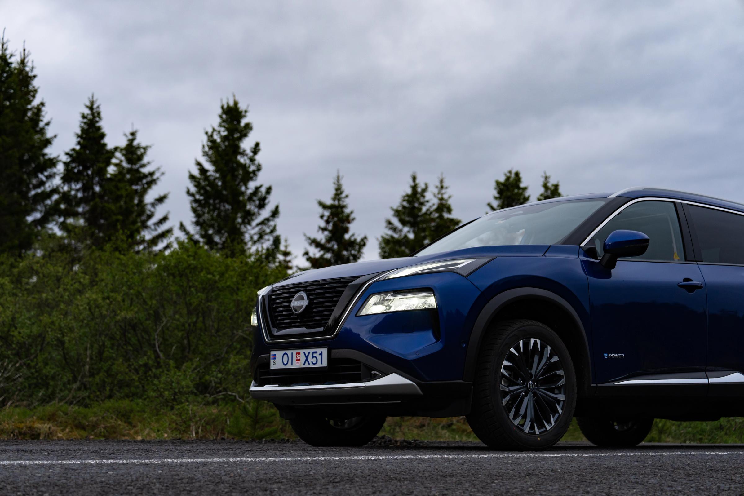 Nissan X-Trail parked beside a paved road with pine trees lining the background