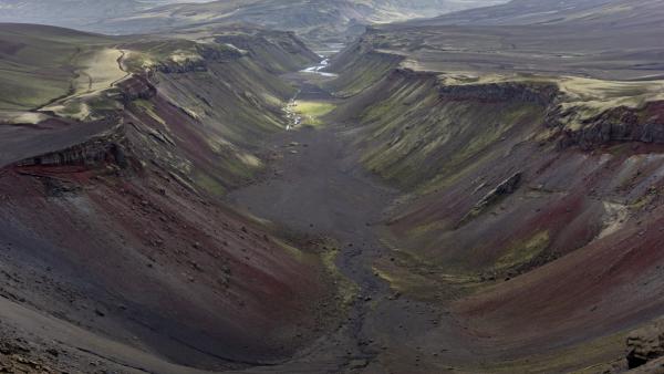 Deep canyon with steep slopes of dark earth, red rock, and green vegetation, and a river at the bottom.