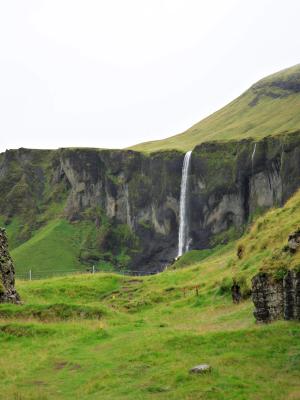 A tall waterfall descends into a green valley between rocky cliffs under an overcast sky.