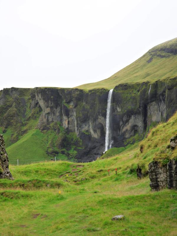 A tall waterfall descends into a green valley between rocky cliffs under an overcast sky.
