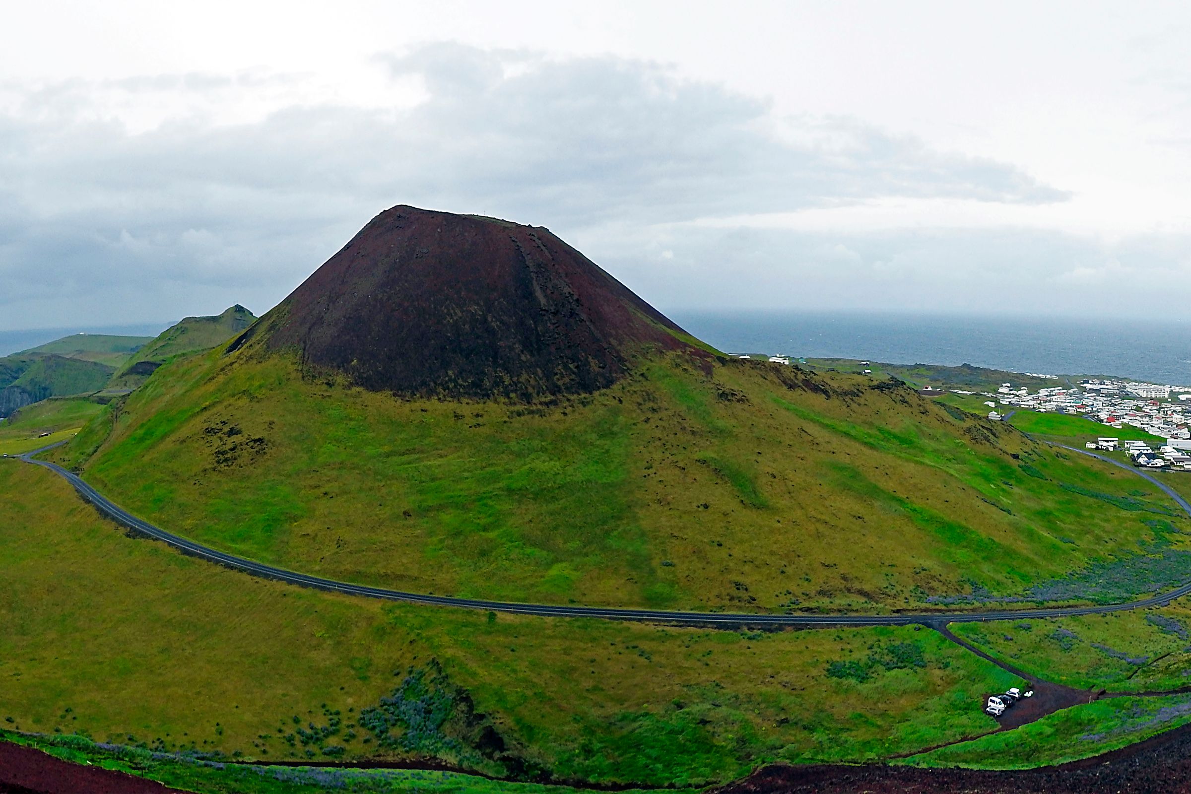 Panoramic view of a volcanic island with a dark-topped volcano, green hills, a coastal town, and the sea under an overcast sky.