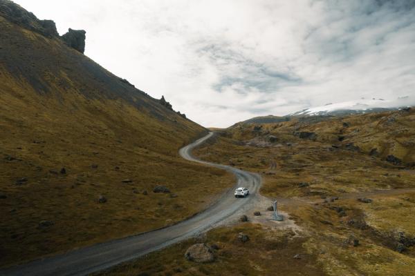 A white car drives on a winding gravel road through a rugged, grassy mountain landscape with a snow-capped peak in the distance under a cloudy sky.