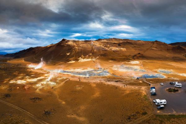 Vast geothermal landscape with steaming vents, orange-brown hills, and a parking area with RVs under a cloudy sky.