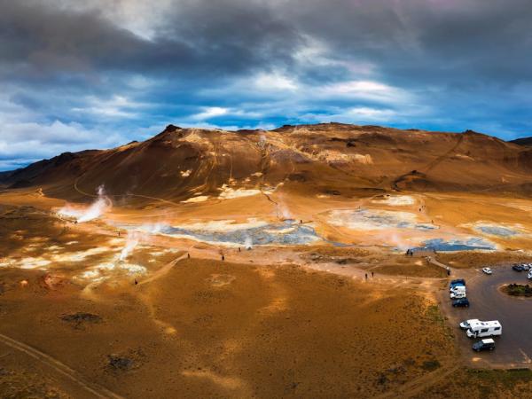 Aerial view of a reddish-brown geothermal landscape with steam vents, discolored ground, a parking lot, and a cloudy sky.