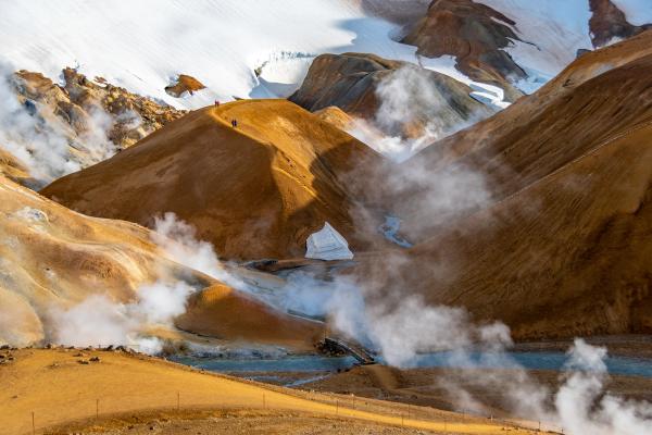 Geothermal landscape with golden-orange hills, snow-capped peaks, a blue river, and white steam rising.