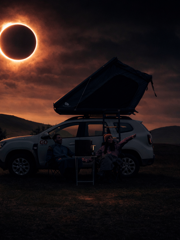 A couple sits beside their car with a rooftop tent, watching a solar eclipse in a dark, cloudy sky.