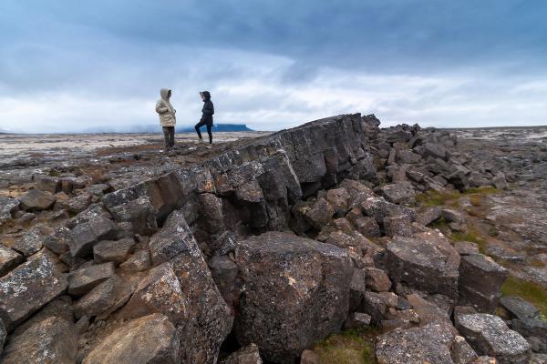 Two people stand on a desolate, rocky plain with columnar rock formations in the foreground under a cloudy sky.