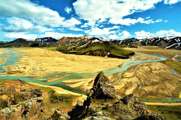 Río trenzado de color turquesa que atraviesa un valle arenoso, rodeado de montañas verdes y nevadas bajo un cielo azul con nubes.