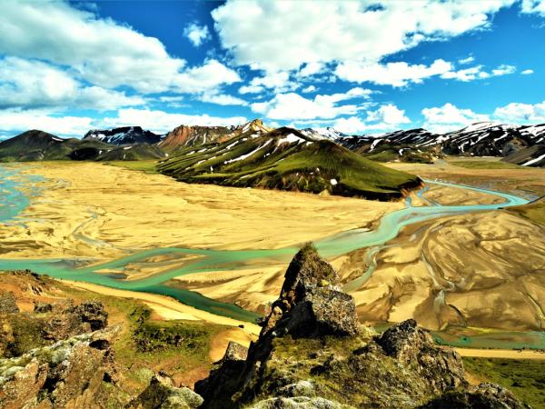 Braided turquoise river winding through a vast sandy valley with green and snow-capped mountains under a blue sky.