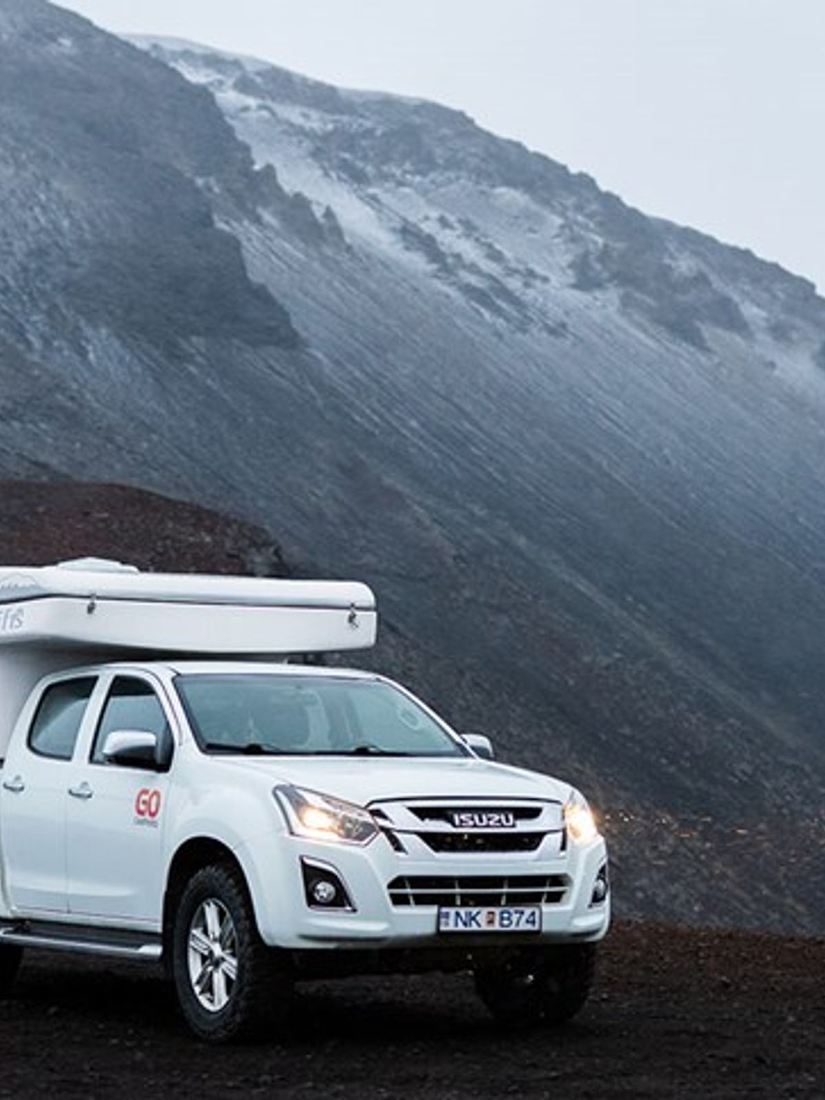 White camper truck on dark, rugged terrain with a snow-capped mountain in the background.